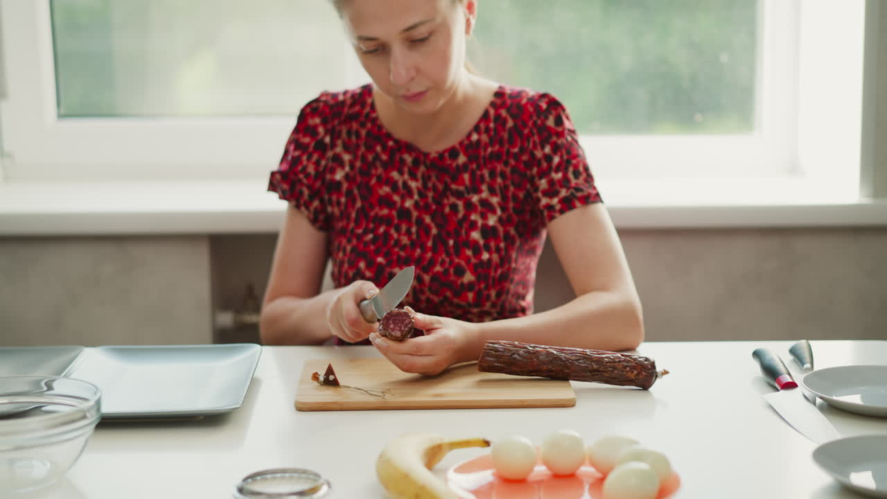 Confectioner in red patterned dress seated by bright window carefully slicing half sausage on wooden board over white table, surrounding plates bowls eggs banana creating serene cooking prep scene