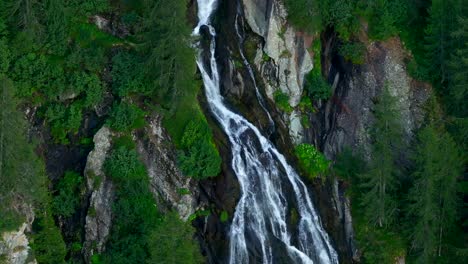 Toma-Aérea-De-Teleobjetivo-De-Una-Estrecha-Cascada-Que-Fluye-Hacia-Abajo-De-La-Montaña,-Alpes-Italianos