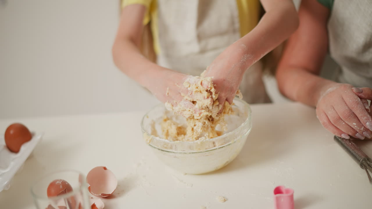 Close up of little kid pulling sticky dough off hands over glass bowl during baking session, flour scattered on white surface, eggshells nearby, partial view of adult offering guidance beside child