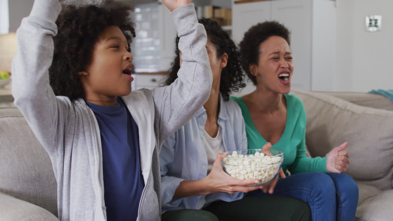 una pareja de lesbianas de raza mixta y su hija viendo la televisión comiendo palomitas de maíz.