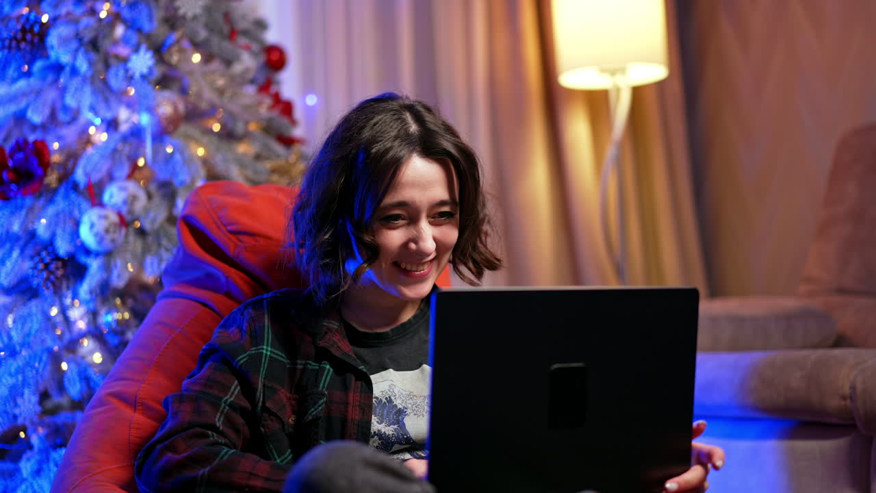 Enjoying a cozy holiday evening at home. A young person sits comfortably in a bean bag chair, joyfully watching something on a laptop during a festive evening