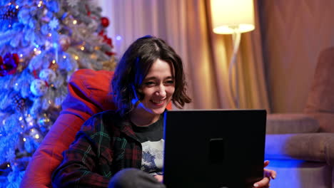 enjoying a cozy holiday evening at home. a young person sits comfortably in a bean bag chair, joyfully watching something on a laptop during a festive evening