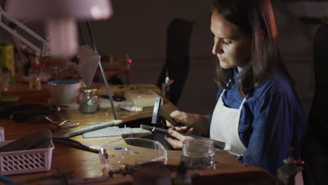 Profile-of-focused-caucasian-female-jeweller-sitting-at-desk,-making-jewelry-in-workshop