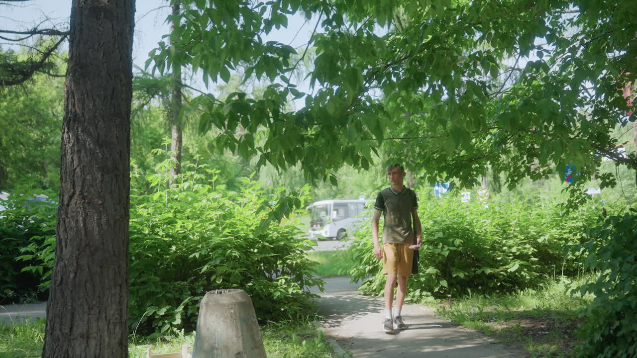 Young White Man Walking Along Shaded Park Path, Cautious Pacing And Scanning Surroundings, Casual Backpack And Summer Shorts, Solitary Approach To Bench Area With Alert Body Language
