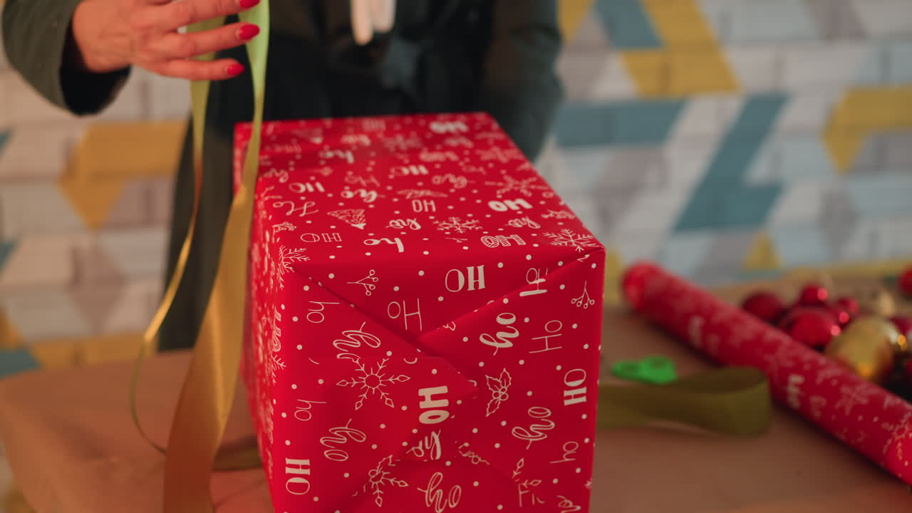 Mani che incartano un regalo di Natale rosso su un tavolo con nastro verde e nastro dorato, ornamenti natalizi e forbici nelle vicinanze, un artigiano fai-da-te lega un fiocco ordinato in un'atmosfera calda e accogliente con uno sfondo a parete fantasia.