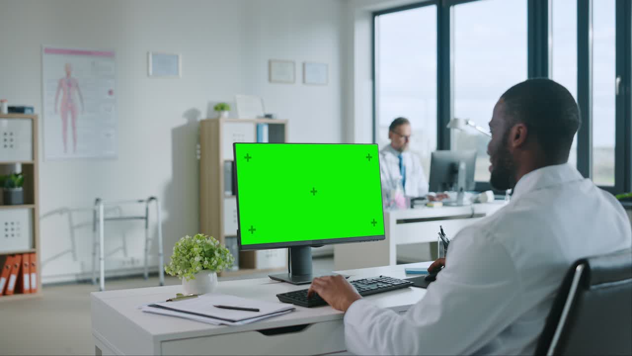 Black Medical Doctor is Working on a Computer with Green Screen Mock Up Display in a Health Clinic. Assistant in White Lab Coat is Reading Medical History Behind a Desk in Hospital Office.