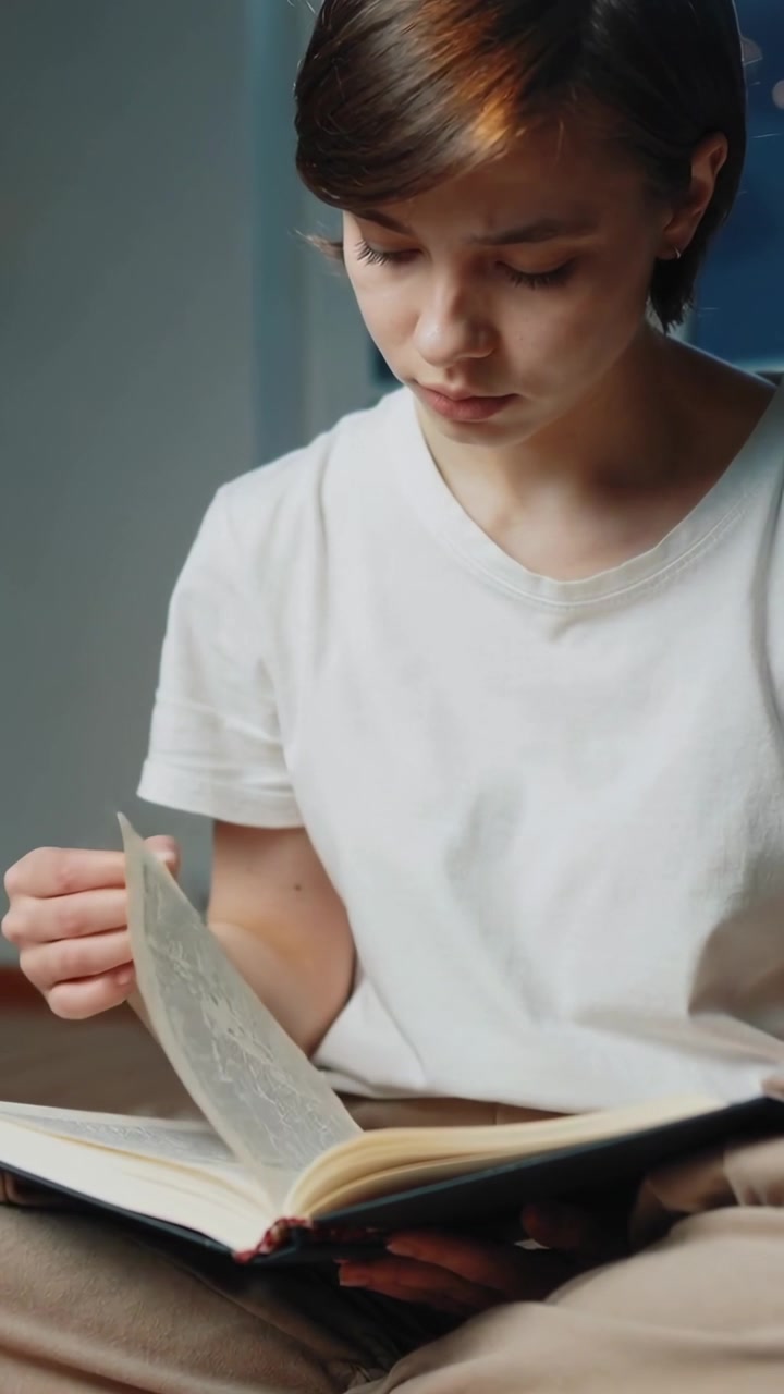 Vertical video: Looking down, teen reading, turning pages holding open book in room in white tee