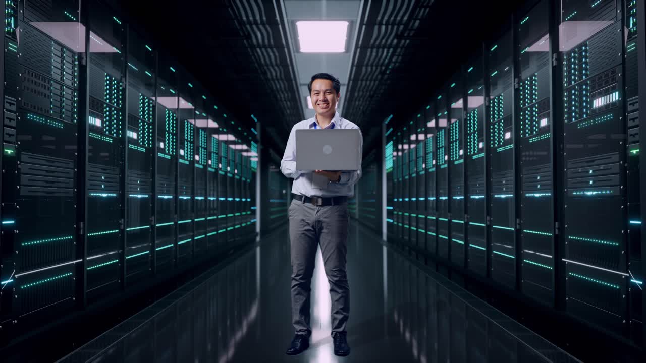 Full Body Of An Asian Male Professional Worker Standing With His Laptop in Server Room Data Center, He Is Looking At The Camera With A Smile