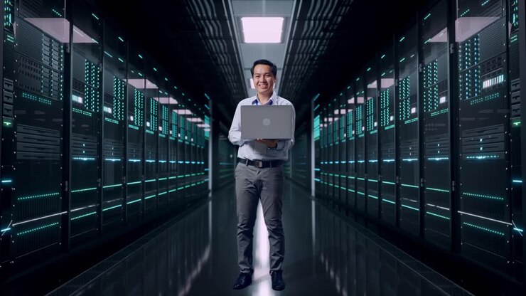 Figura intera di un lavoratore professionista asiatico maschio in piedi con il suo laptop nella sala server di un data center, guarda la fotocamera con un sorriso