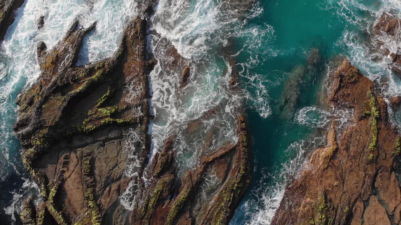 Inquadratura statica dall'alto delle acque di Porto Das Salemas, con le loro acque turchesi che si infrangono sulla roccia marrone schizzando schiuma bianca al centro della vista