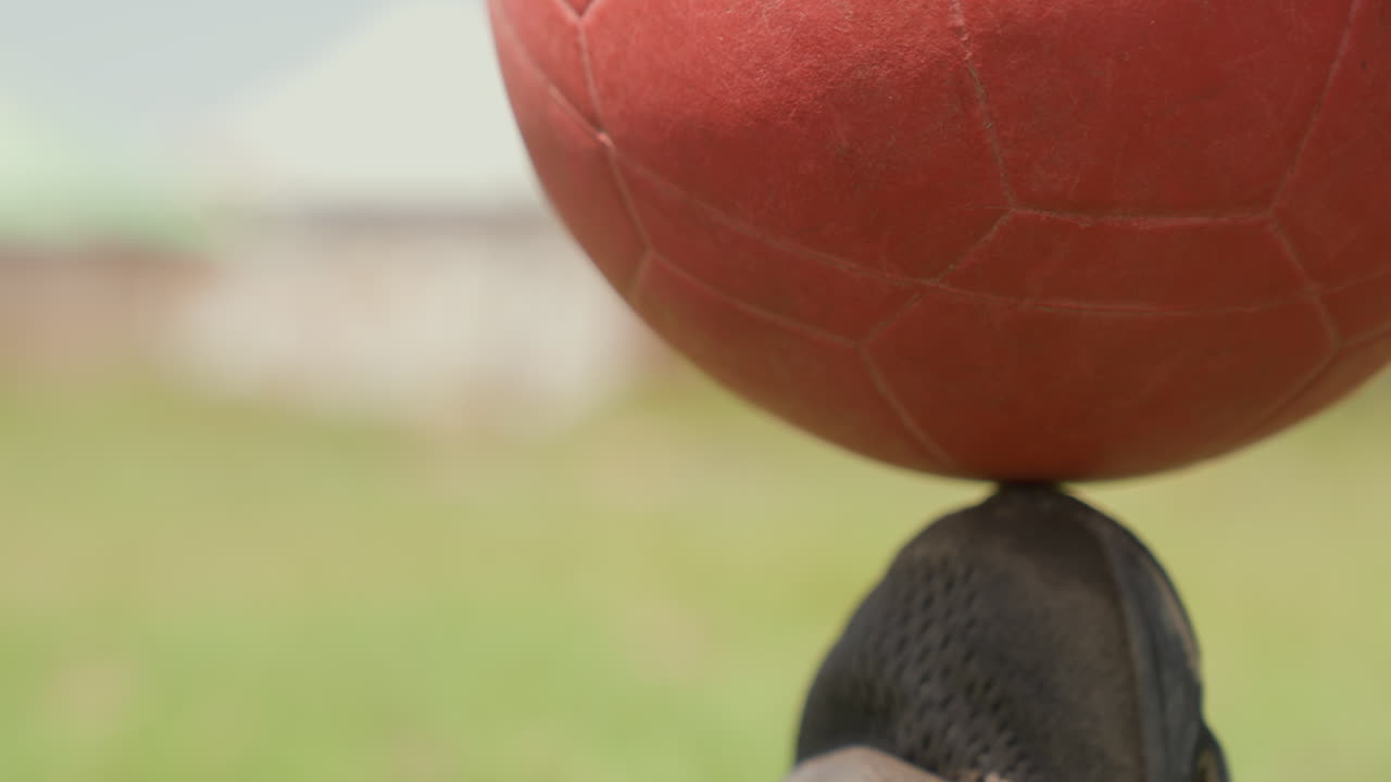 Athlète noir équilibrant un ballon sur sa chaussure, gros plan sur l'entraînement de précision de la pointe de la basket, répétition disciplinée sur un terrain ensoleillé, cadrage serré sur la technique et le contrôle du mouvement, concentration intense.