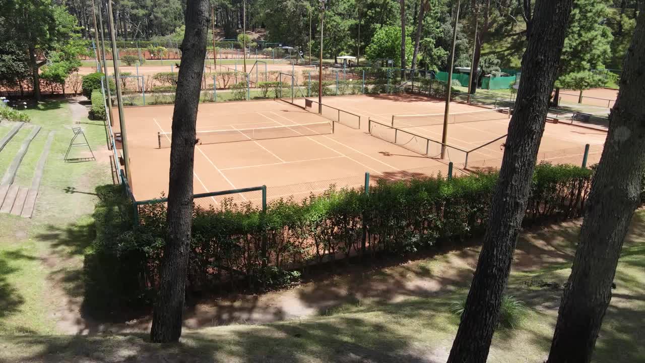 Multiple empty tennis clay courts are shown from a high vantage point, surrounded by lush green trees