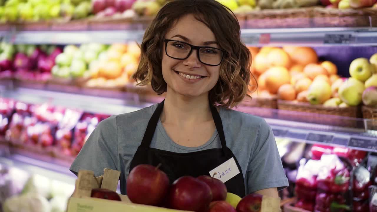 Beautiful smiling young female supermarket employee in black apron holding a box full of apples in front of shelf in supermarket