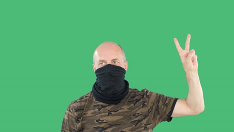 bald man in military shirt and face bandana showing two fingers on green chromakey background. protester man showing victory fingers on green background.