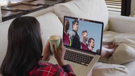 Composite-of-woman-sitting-at-home-holding-coffee-watching-football-match-on-laptop