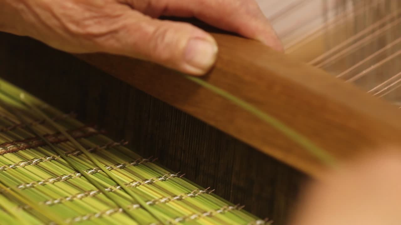 Detailed view of a weaving process using green plant stalks and thin threads on a wooden loom. A hand gently guides the material through the pattern
