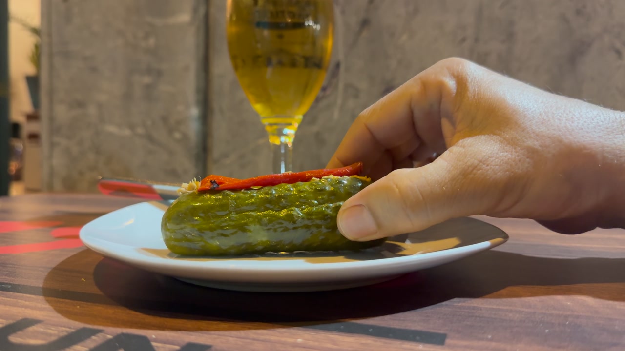 Hand of a caucasian man wearing bracelets eating pickle filled with tuna fish and drinking a cup of beer in a bar.