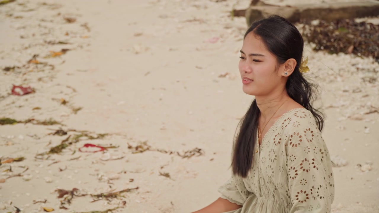 Wanita Asia minum dari cangkir di pantai, duduk di pasir dekat rumput laut, ekspresi close-up, rambut hitam panjang, gaun linen bermotif bunga, gerak-gerik penuh pemikiran, napas perlahan, suara ombak yang lembut, potret candid.