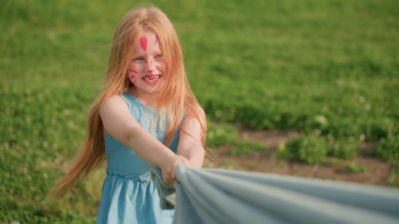 Cat face painted girl dragging blanket across grass while smiling, sunlight catching hair, playful energy in summer park, family figures blurred in background, joyful outdoor childhood moment