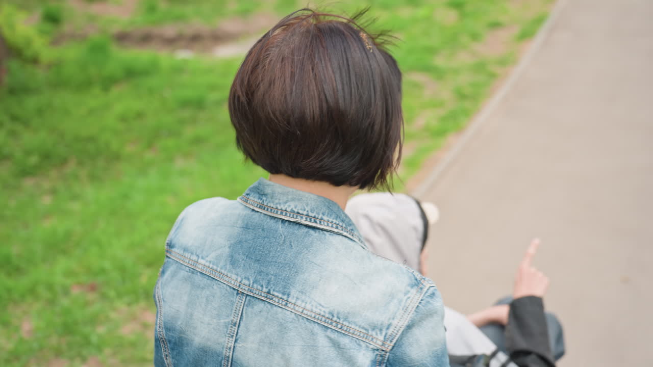 Rear View Of Caregiver And Child On Park Path, Short Haircut And Denim Jacket Visible, Peaceful Stroll With Attentive Presence And Green Trees Lining Sidewalk, Gentle Supportive Scene