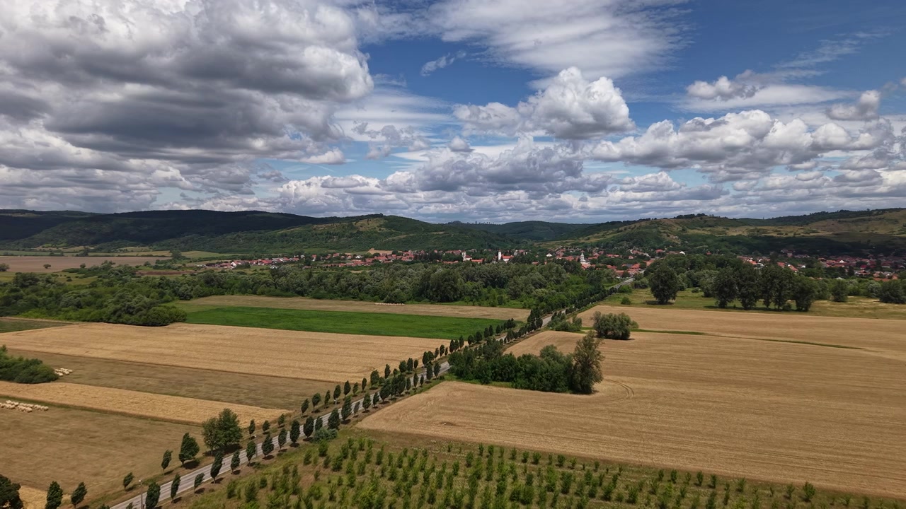 Vista aerea che cattura campi dorati, una strada tortuosa e un incantevole villaggio annidato tra verdi colline sotto un cielo nuvoloso e drammatico.
