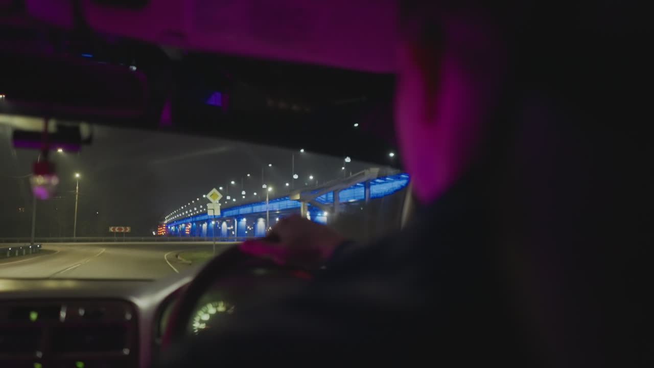 aproximación nocturna al puente desde el asiento del conductor, interior bañado en neón azul, instrumentos del salpicadero iluminados, tramo iluminado por delante, conductor centrado en la silueta navegando por la autopista urbana