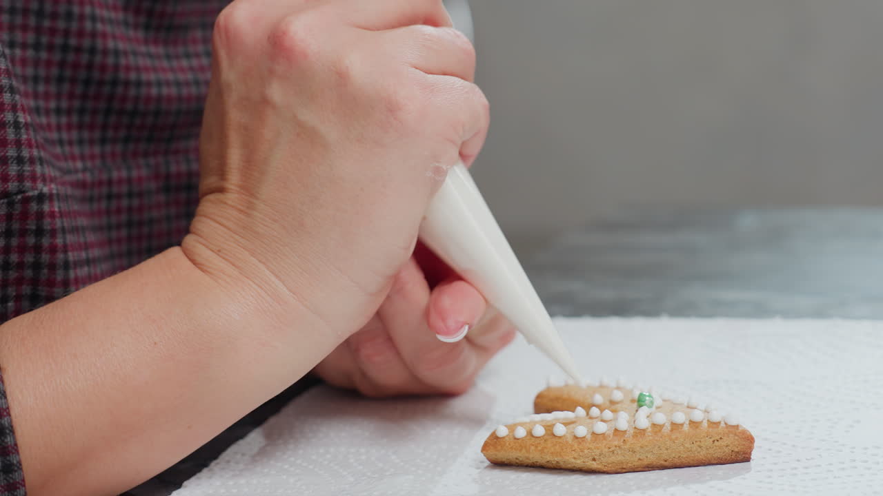 색 아이싱 점과 녹색 구슬을 가진 사랑 모양의 쿠키를 장식하는 손의 측면 보기, 쿠키는 색 텍스처 된 종이에 배치되며 아이싱은 파이핑 가방을 사용하여 조심스럽게 적용됩니다.