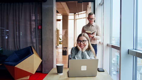 positive smiling blonde looks at her laptop sitting at desk. man stands with a book and coffee cup in hands at the window at backdrop.