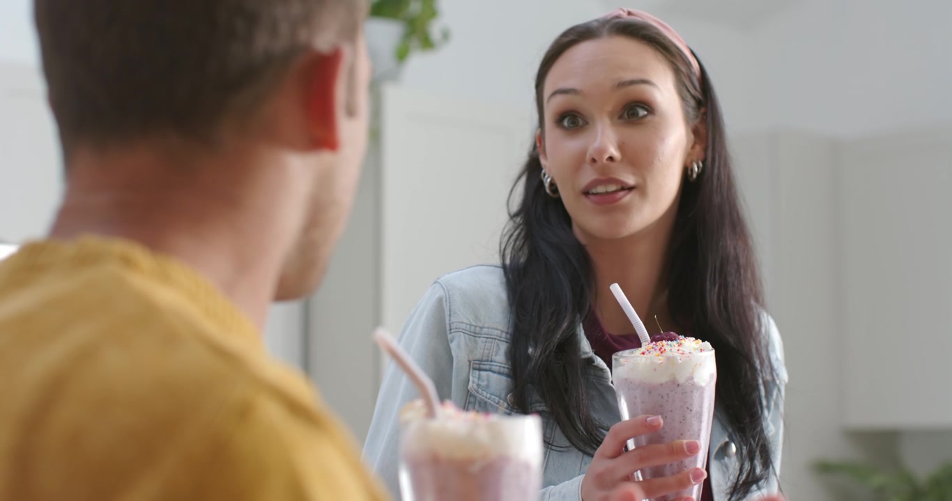 Woman standing at counter holding milkshake, sipping and replying to man gesturing