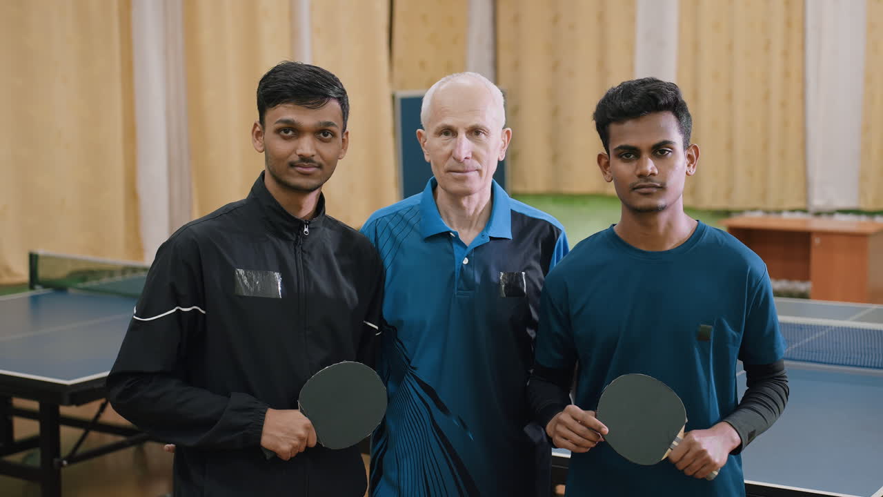 Coach stands proudly with two young players holding table tennis rackets during indoor photoshoot, symbolizing unity, mentorship, teamwork, training, confidence, and dedication