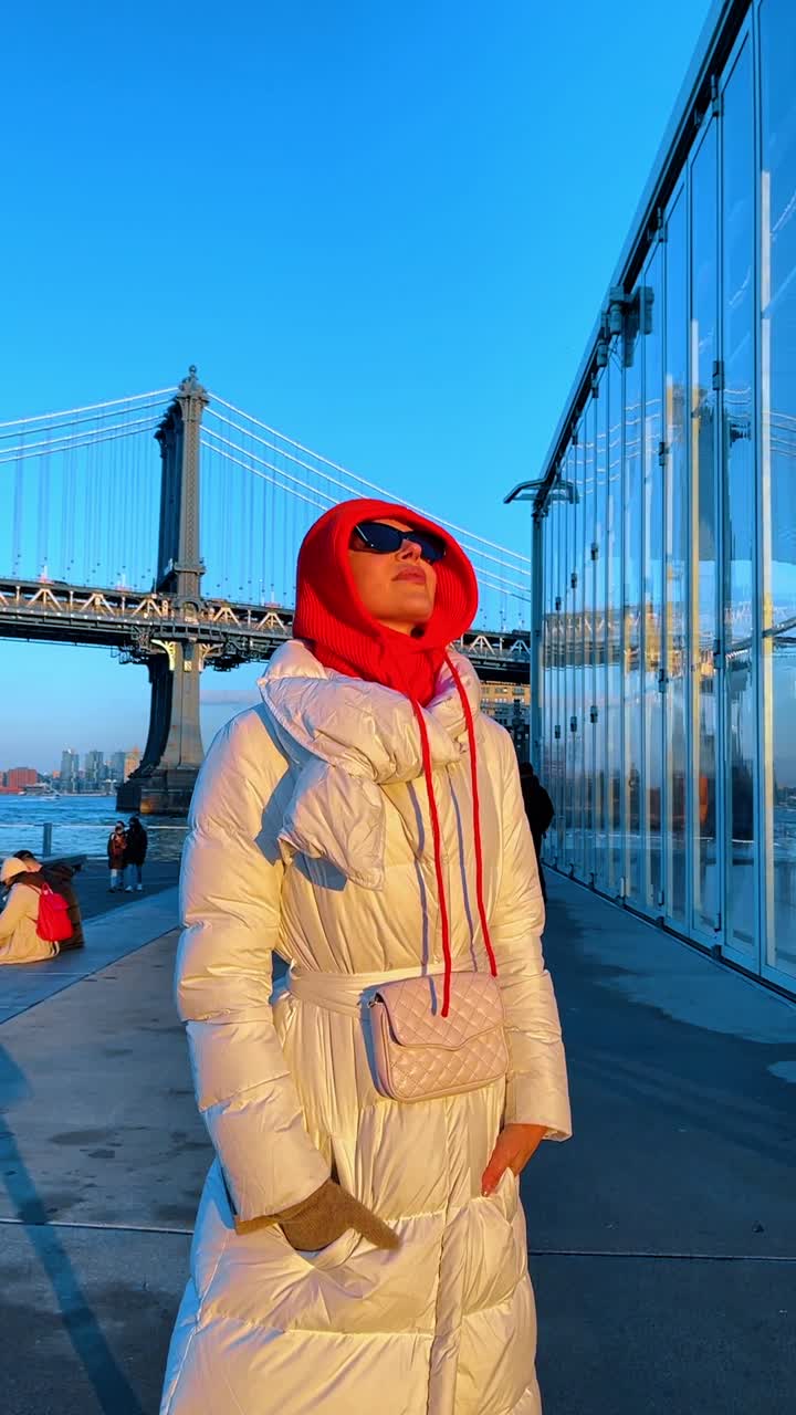 New York, USA, 2 novembre 2022: Donna in cappotto bianco vicino al Ponte di Manhattan. Donna con cappotto piumino bianco e cappuccio rosso in piedi vicino al Ponte di Manhattan, lungomare di Brooklyn, luce dorata.