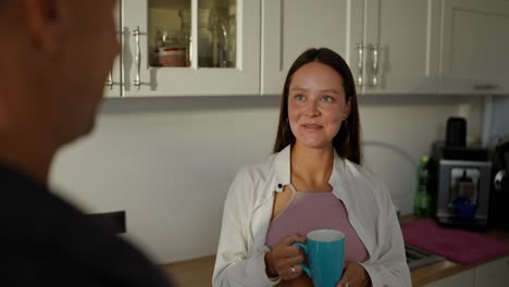 Over-the-shoulder-a-happy-pregnant-brunette-girl-in-a-white-shirt-drinks-tea-from-a-blue-cup-and-talks-with-her-husband-in-the-morning-in-the-kitchen