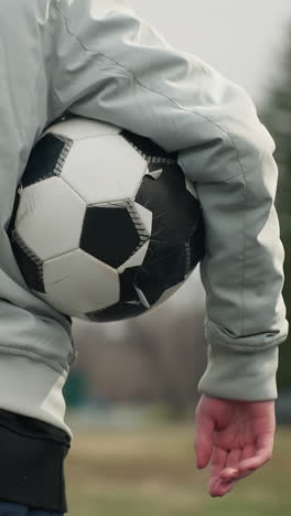 back view of a person standing with a soccer ball tucked under the right arm, wearing a gray jacket with their left hand in the pocket, the background shows a blurred view of a grassy field