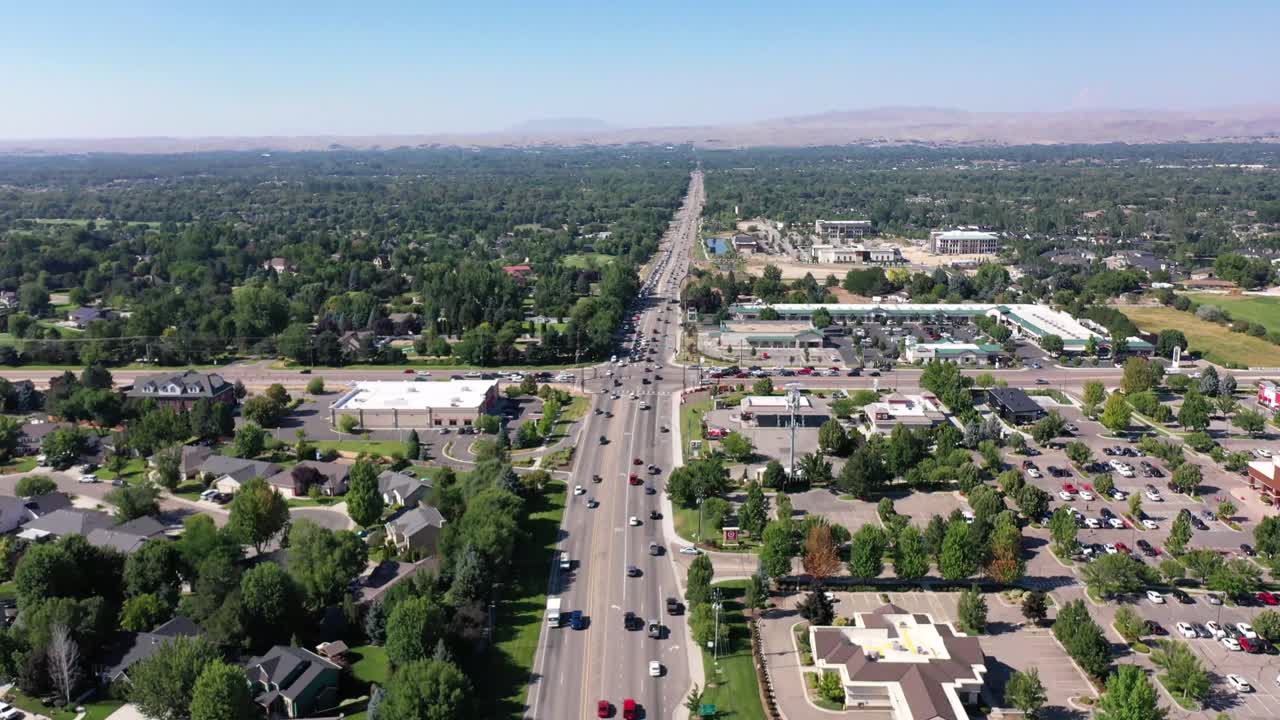 Drone pushes in and tilts down over a steady flow of traffic. There are lots of visible trees, homes and businesses.