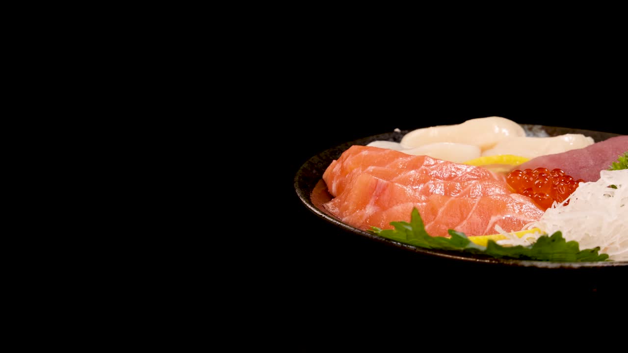 A plate of assorted sashimi, including salmon, tuna, scallop, and salmon roe, rotates smoothly under studio lighting against a seamless black backdrop