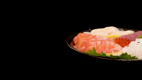 a plate of assorted sashimi, including salmon, tuna, scallop, and salmon roe, rotates smoothly under studio lighting against a seamless black backdrop