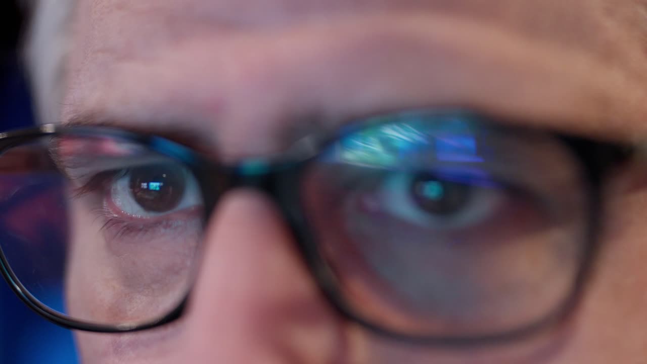 Primo piano di un uomo concentrato con gli occhiali che riflettono la luce blu di un monitor di computer, a indicare il lavoro su un progetto digitale. L'espressione trasmette serietà e attenzione