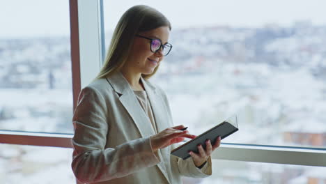 young woman taking notes in her book standing at the window. lady in light jacket looks thoughtfully into the blurred window.