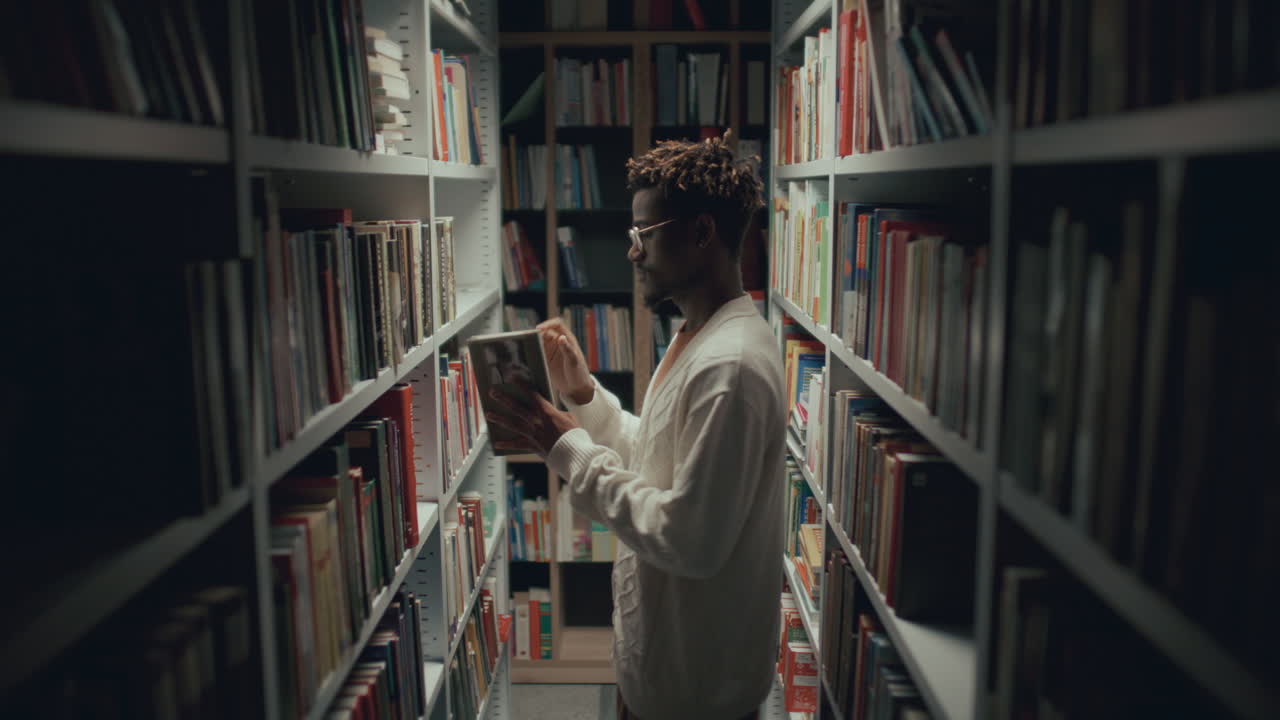 hombre afroamericano de pie en el pasillo de la biblioteca, recogiendo un libro de la estantería