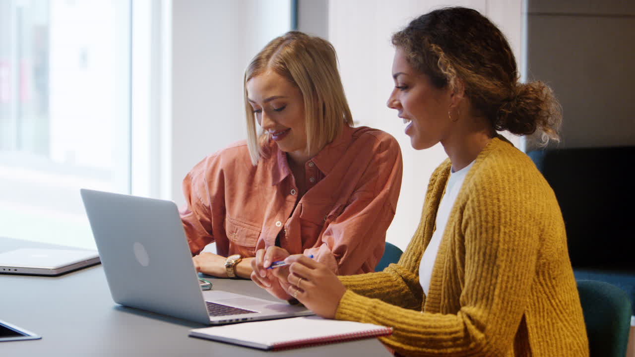 dos jóvenes mujeres creativas sentadas en una oficina trabajando juntas en una computadora portátil, de cerca