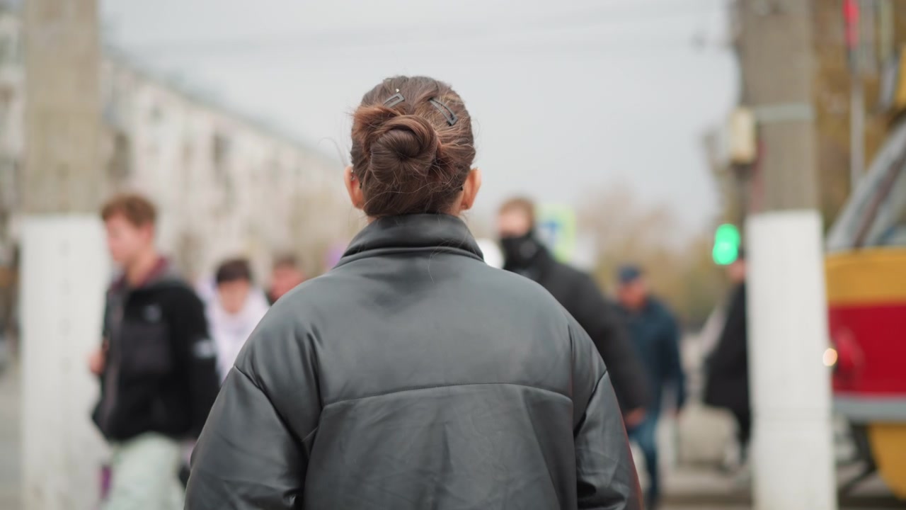 Primo piano di donna con capelli castani raccolti a chignon, occhiali e giacca di pelle nera, che cammina per una trafficata strada cittadina. Lo sfondo mostra pedoni, edifici, alberi e veicoli sfocati.