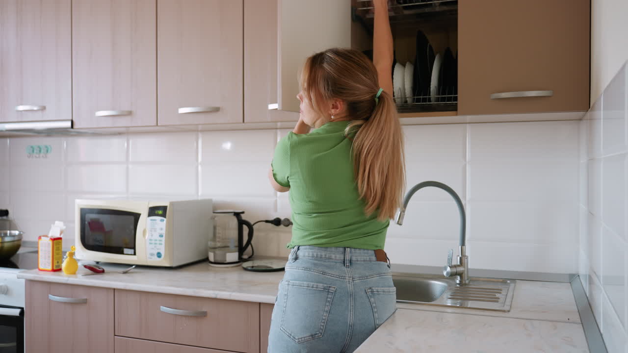 Visuale posteriore di una donna che riordina una cucina moderna mentre ripone una tazza nera in un pensile, mantenendo l'ordine tra forno a microonde, lavandino, fornello e utensili durante la sua routine quotidiana.