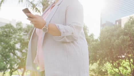 plus size biracial woman walking with luggage and using smartphone in city