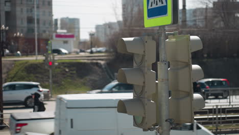 close view of a city traffic light and pedestrian crossing sign in an urban area, with cars passing by and someone standing by the road pavement