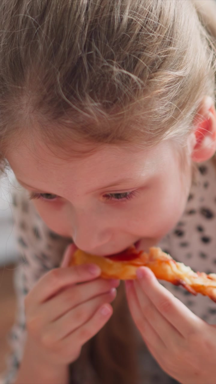 Close up of young girl eating pizza slice holding food with both hands throughout. Pizza crust and toppings clearly visible as girl brings it to mouth. Soft indoor lighting with blurred