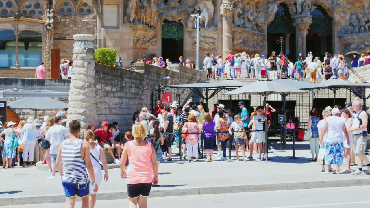 많은 관광객들이 바르셀로의 사그라다 파밀리아 교회 입구에서 걷고 사진을 찍습니다.