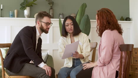 Young-Woman-Holding-Paper-Report-And-Debating-With-Her-Two-Colleagues-While-Sitting-In-Wooden-Chairs-In-A-Living-Room-1