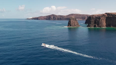 Roque-Partido,-Gran-Canaria:-Vista-Aérea-En-órbita-Y-Seguimiento-Lateral-Sobre-El-Barco-Y-Al-Fondo-El-Majestuoso-Roque-Partido-En-Un-Día-Soleado