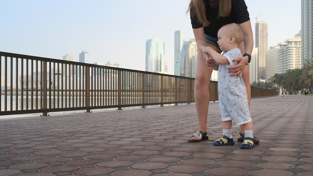 un niño sonriente sosteniendo la mano de su madre hace los primeros pasos caminando por el paseo marítimo en verano.