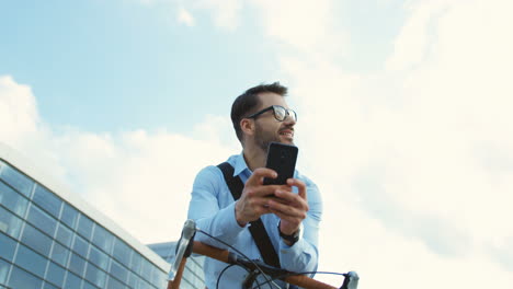 View-from-below-of-cheerful-man-wearing-glasses-and-leaning-on--his-bike,-holding-smartphone-and-looking-at-the-sides-with-a-smile